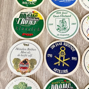 Pu&ograve; includere: Una collezione di sottobicchieri rotondi in cartone con vari disegni grafici, tra cui il marchio Heineken, testi commemorativi, illustrazioni di navi, simboli di artiglieria e bicchieri di birra con dettagli stampati a colori.