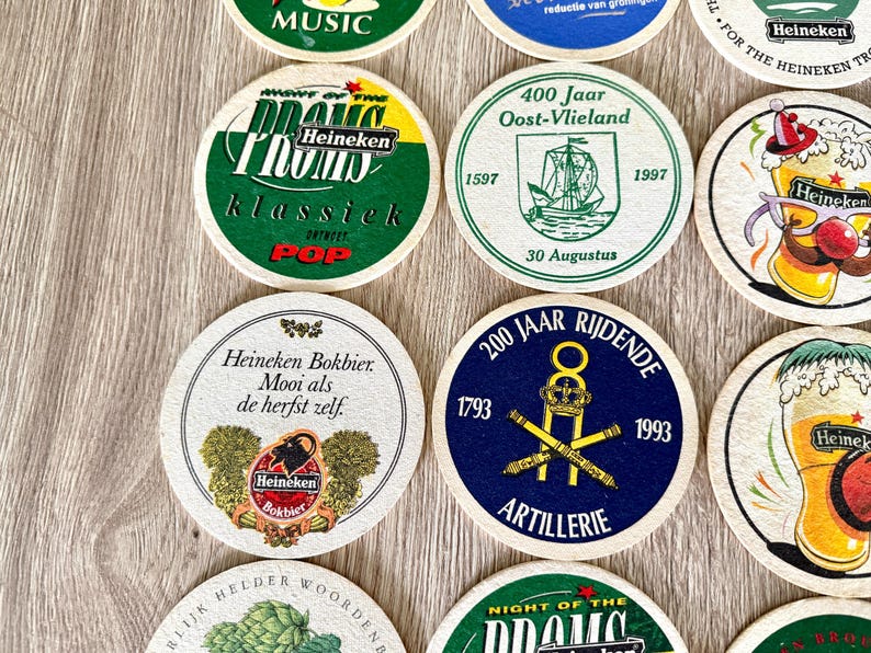 Pu&ograve; includere: Una collezione di sottobicchieri rotondi in cartone con vari disegni grafici, tra cui il marchio Heineken, testi commemorativi, illustrazioni di navi, simboli di artiglieria e bicchieri di birra con dettagli stampati a colori.