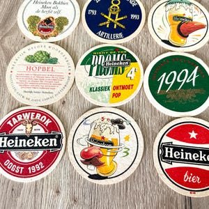 Pu&ograve; includere: Una collezione di sottobicchieri rotondi in cartone con vari marchi Heineken, inclusi loghi, date come 1992 e 1994, e illustrazioni grafiche di bicchieri di birra, luppolo e testi come Night of the Proms e Hopbel.