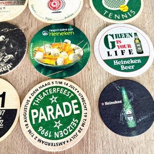 Pu&ograve; includere: Una collezione di sottobicchieri circolari in cartone con vari design, tra cui il marchio Heineken, una grafica da tennis, una pubblicit&agrave; per un festival jazz e un annuncio per una parata teatrale, tutti disposti in una griglia.