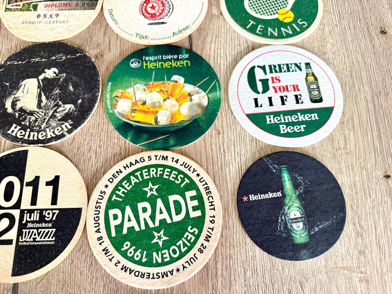Pu&ograve; includere: Una collezione di sottobicchieri circolari in cartone con vari design, tra cui il marchio Heineken, una grafica da tennis, una pubblicit&agrave; per un festival jazz e un annuncio per una parata teatrale, tutti disposti in una griglia.