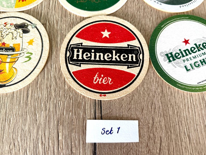 Pu&ograve; includere: Una collezione di sottobicchieri circolari in cartone con vari design del marchio Heineken. Il sottobicchiere centrale mostra una grafica rossa e bianca con il testo Heineken bier e una piccola stella. Un'etichetta bianca rettangolare riporta Set 1.