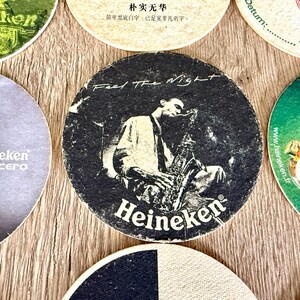 Pu&ograve; includere: Un sottobicchiere rotondo in cartone con una grafica in bianco e nero di una persona che suona il sassofono. Il testo "Feel The Night" &egrave; stampato in alto e il logo "Heineken" &egrave; stampato in bianco in basso su uno sfondo scuro e strutturato.