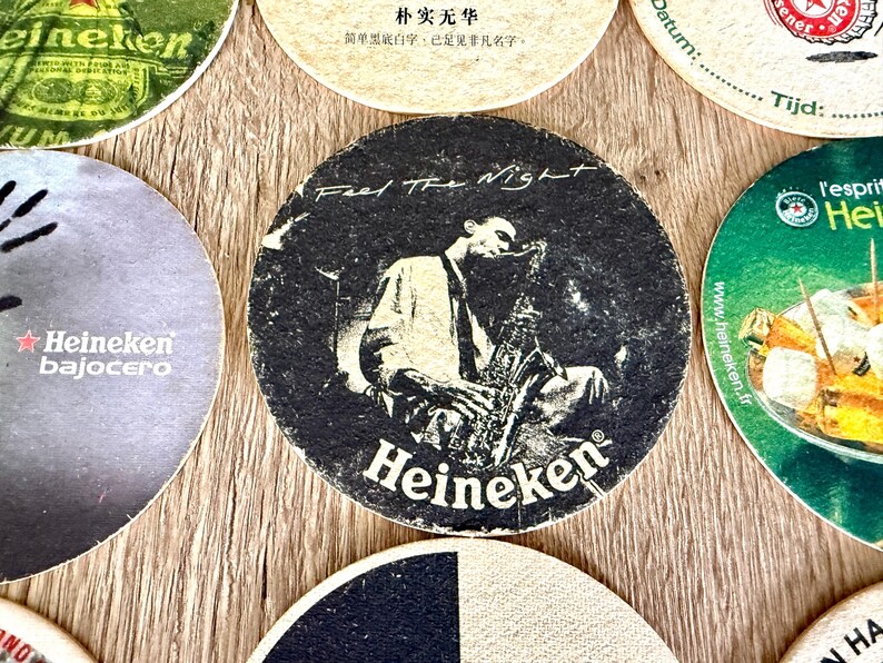 Pu&ograve; includere: Un sottobicchiere rotondo in cartone con una grafica in bianco e nero di una persona che suona il sassofono. Il testo "Feel The Night" &egrave; stampato in alto e il logo "Heineken" &egrave; stampato in bianco in basso su uno sfondo scuro e strutturato.