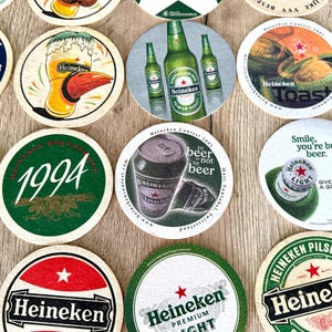 Pu&ograve; includere: Una collezione di sottobicchieri circolari in cartone con vari marchi Heineken, inclusi grafiche di bottiglie, lattine e bicchieri di birra, con testo stampato come 1994, Toast e to beer or not to beer, disposti su una superficie piana.