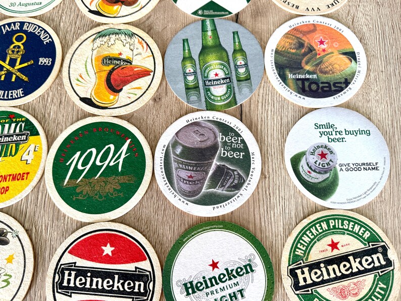 Pu&ograve; includere: Una collezione di sottobicchieri circolari in cartone con vari marchi Heineken, inclusi grafiche di bottiglie, lattine e bicchieri di birra, con testo stampato come 1994, Toast e to beer or not to beer, disposti su una superficie piana.