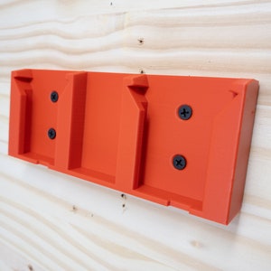 Puede incluir: Soporte de pared de pl&aacute;stico naranja con tres ranuras para sujetar objetos. El soporte tiene cuatro tornillos negros.