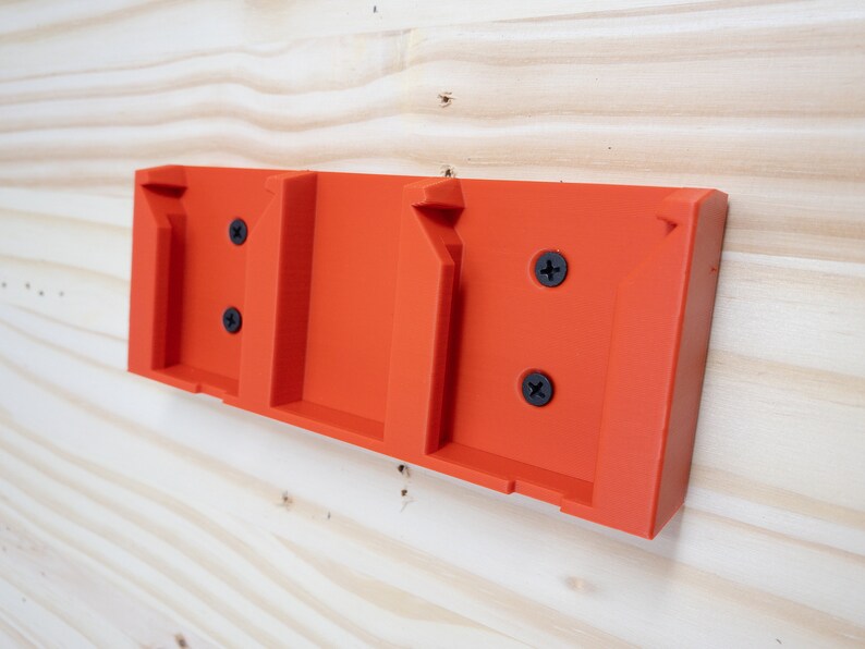 Puede incluir: Soporte de pared de pl&aacute;stico naranja con tres ranuras para sujetar objetos. El soporte tiene cuatro tornillos negros.