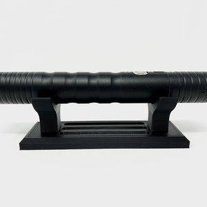 Puede incluir: Una r&eacute;plica de sable l&aacute;ser negro en un soporte negro. El sable l&aacute;ser tiene una superficie texturizada y un bot&oacute;n en el lateral.