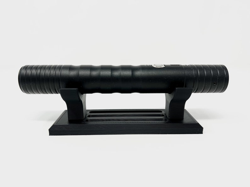 Puede incluir: Una r&eacute;plica de sable l&aacute;ser negro en un soporte negro. El sable l&aacute;ser tiene una superficie texturizada y un bot&oacute;n en el lateral.