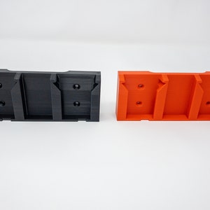 Puede incluir: Dos soportes de pared impresos en 3D, uno negro y otro naranja, con tres ranuras para sujetar objetos.