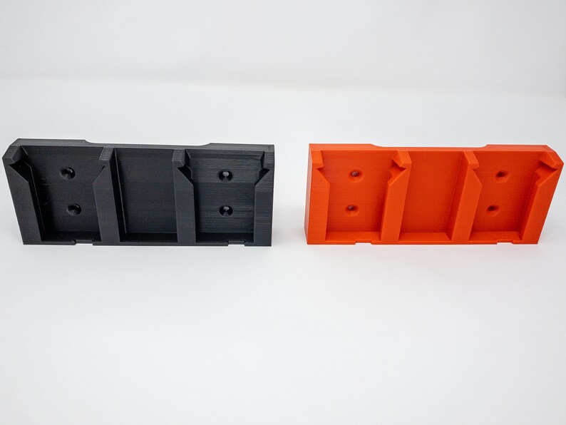 Puede incluir: Dos soportes de pared impresos en 3D, uno negro y otro naranja, con tres ranuras para sujetar objetos.