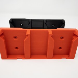 Puede incluir: Un soporte de pared de pl&aacute;stico naranja impreso en 3D con tres ranuras y dos agujeros a cada lado. El soporte est&aacute; apilado sobre un soporte de pared de pl&aacute;stico negro similar.