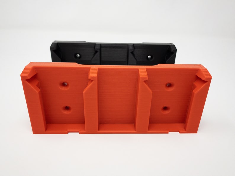 Puede incluir: Un soporte de pared de pl&aacute;stico naranja impreso en 3D con tres ranuras y dos agujeros a cada lado. El soporte est&aacute; apilado sobre un soporte de pared de pl&aacute;stico negro similar.