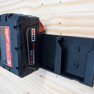 Puede incluir: Una bater&iacute;a Milwaukee M18 REDLITHIUM XC3.0 negra y roja est&aacute; montada en un soporte de pared de pl&aacute;stico negro. El soporte de pared tiene dos tornillos.