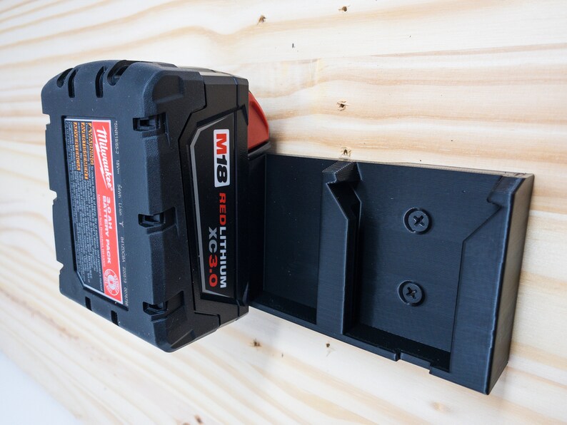 Puede incluir: Una bater&iacute;a Milwaukee M18 REDLITHIUM XC3.0 negra y roja est&aacute; montada en un soporte de pared de pl&aacute;stico negro. El soporte de pared tiene dos tornillos.
