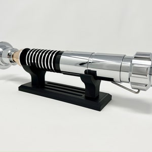 Puede incluir: Un sable de luz cromado con detalles negros en el mango se muestra en un soporte negro. El sable de luz tiene un detalle dorado cerca del mango.