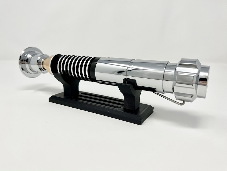 Puede incluir: Un sable de luz cromado con detalles negros en el mango se muestra en un soporte negro. El sable de luz tiene un detalle dorado cerca del mango.