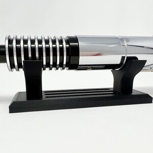 Puede incluir: Un sable de luz cromado con un mango negro y un acento dorado en el extremo. El sable de luz est&aacute; en un soporte negro.