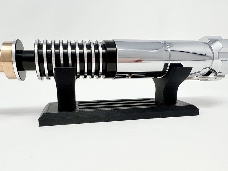 Puede incluir: Un sable de luz cromado con un mango negro y un acento dorado en el extremo. El sable de luz est&aacute; en un soporte negro.