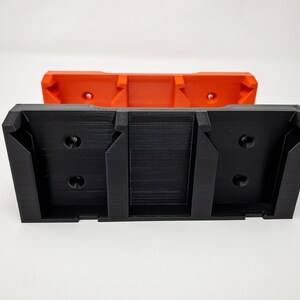 Puede incluir: Un soporte de pared de impresi&oacute;n 3D negro con dos ranuras para sujetar objetos. El soporte est&aacute; unido a una pieza m&aacute;s peque&ntilde;a de impresi&oacute;n 3D naranja.