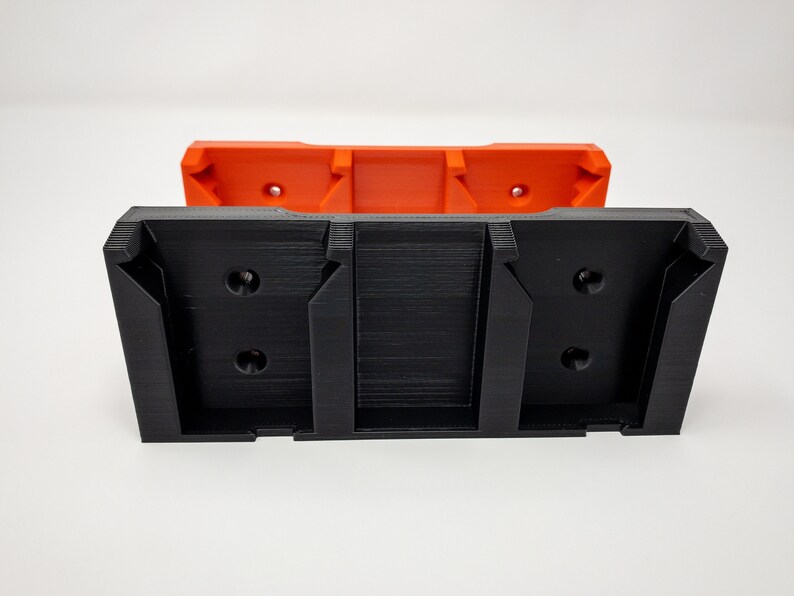 Puede incluir: Un soporte de pared de impresi&oacute;n 3D negro con dos ranuras para sujetar objetos. El soporte est&aacute; unido a una pieza m&aacute;s peque&ntilde;a de impresi&oacute;n 3D naranja.