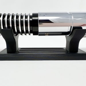 Puede incluir: Una r&eacute;plica de sable l&aacute;ser negro con una hoja plateada y un soporte negro. El sable l&aacute;ser est&aacute; en un soporte negro con una hoja plateada.