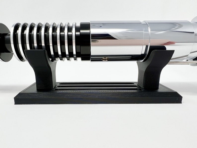 Puede incluir: Una r&eacute;plica de sable l&aacute;ser negro con una hoja plateada y un soporte negro. El sable l&aacute;ser est&aacute; en un soporte negro con una hoja plateada.