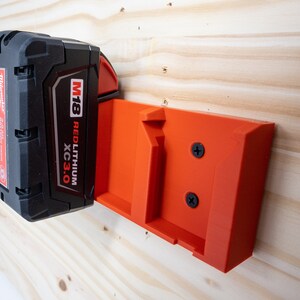 Puede incluir: Un soporte de pared de pl&aacute;stico rojo para una bater&iacute;a Milwaukee M18 Red Lithium XC 3.0. El soporte est&aacute; fijado a una superficie de madera con dos tornillos.