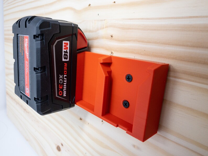 Puede incluir: Un soporte de pared de pl&aacute;stico rojo para una bater&iacute;a Milwaukee M18 Red Lithium XC 3.0. El soporte est&aacute; fijado a una superficie de madera con dos tornillos.