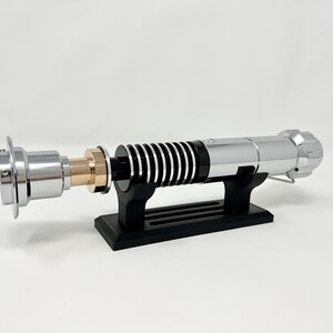 Puede incluir: Un sable de luz plateado con un mango negro y plateado, sobre un soporte negro. El sable de luz tiene un acento dorado en el mango.