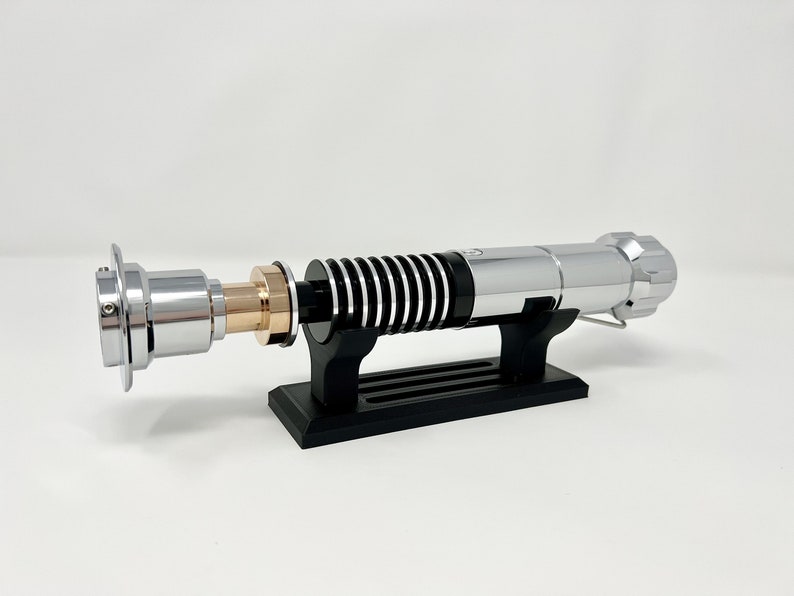 Puede incluir: Un sable de luz plateado con un mango negro y plateado, sobre un soporte negro. El sable de luz tiene un acento dorado en el mango.