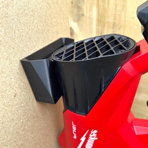May include: A red and black power tool with a black accessory attached to a wooden surface. The tool has the brand name "Milwaukee" printed on it in white. The accessory is a black, angled holder.