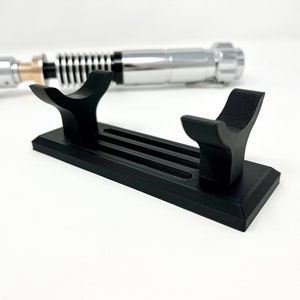 Puede incluir: Un soporte de sable de luz de pl&aacute;stico negro con dos ranuras para sostener sables de luz. El soporte tiene una base rectangular y dos brazos curvos.