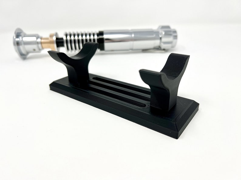 Puede incluir: Un soporte de sable de luz de pl&aacute;stico negro con dos ranuras para sostener sables de luz. El soporte tiene una base rectangular y dos brazos curvos.