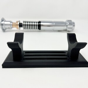 Puede incluir: Un soporte de sable de luz de pl&aacute;stico negro con dos brazos curvos y una base rectangular con cinco ranuras. El soporte est&aacute; dise&ntilde;ado para sostener un sable de luz en posici&oacute;n vertical.