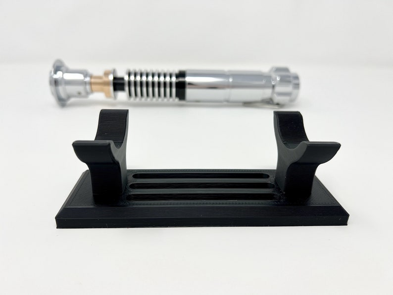 Puede incluir: Un soporte de sable de luz de pl&aacute;stico negro con dos brazos curvos y una base rectangular con cinco ranuras. El soporte est&aacute; dise&ntilde;ado para sostener un sable de luz en posici&oacute;n vertical.