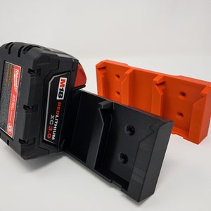 Puede incluir: Un soporte de bater&iacute;a negro y naranja con una bater&iacute;a Milwaukee M18 REDLITHIUM XC3.0 conectada. El soporte est&aacute; dise&ntilde;ado para montarse en una pared u otra superficie.