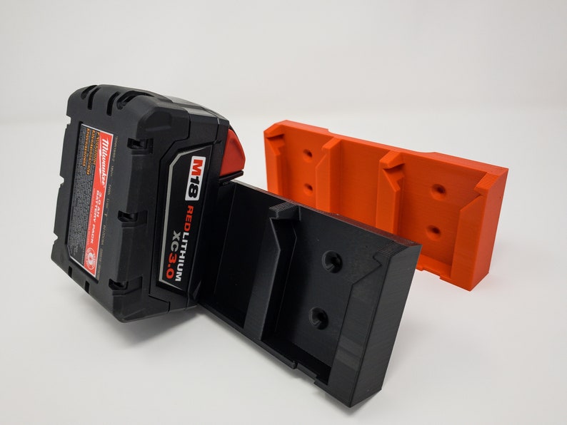 Puede incluir: Un soporte de bater&iacute;a negro y naranja con una bater&iacute;a Milwaukee M18 REDLITHIUM XC3.0 conectada. El soporte est&aacute; dise&ntilde;ado para montarse en una pared u otra superficie.