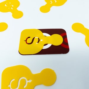 May include: A yellow die-cut shape with a dollar sign cutout is placed over a maroon and white business card. Several other yellow shapes with dollar sign cutouts are scattered around the card on a white background.