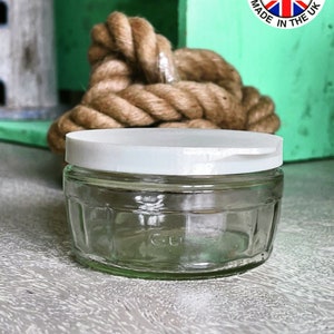 May include: A clear glass jar with a white lid. The jar has a textured surface and is sitting on a light wood surface.