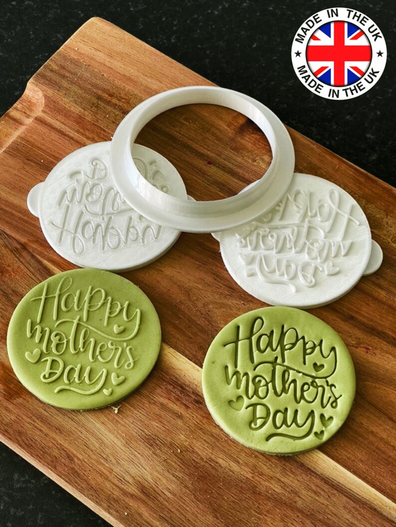 May include: Two green fondant cookies with the text "Happy Mother's Day" in white script. The cookies are on a wooden surface with three white plastic cookie cutters. One cutter is a circle, and the other two are round with the text "Happy Mother's Day" in white script.