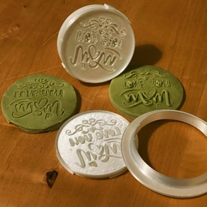 May include: Four cookie cutters, two are silver and two are green. The silver cutters are engraved with the words "New Mom" and "Love You Mom". The green cutters are imprinted with the same text. The cutters are on a wooden surface.