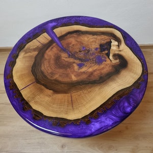 May include: Round wooden table with a natural wood grain center and a vibrant purple resin border. The table's surface features a unique design with visible wood rings and a glossy finish, showcasing a blend of natural and modern elements.