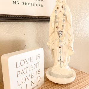 May include: A white statue of a woman with a rosary. The rosary has gray and brown beads and a silver cross. A white block with the text "LOVE IS PATIENT LOVE IS KIND 1 CORINTHIANS 13:4" is on the left. A framed print with the text "MY LORD. MY SHEPHERD." is in the background.