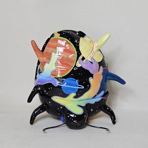 May include: A whimsical, hand-painted ceramic sculpture of a beetle-like creature with a black body and colorful space-themed details. The sculpture features planets, stars, and a yellow horn-like element. The legs and antennae are black.