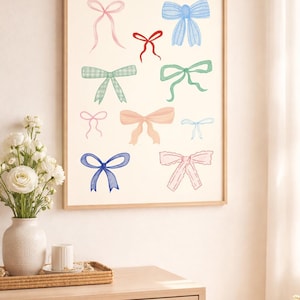 May include: Watercolor print of various bows in shades of pink, blue, green, and peach. The bows are arranged on a cream-colored background and framed in a light wood frame. The artwork is displayed on a light wood dresser.