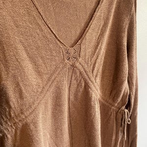 Puede incluir: Una blusa de punto marr&oacute;n con cuello en V y mangas largas. La blusa presenta un detalle calado decorativo en el escote y lazos en los laterales. La tela tiene un aspecto texturizado.