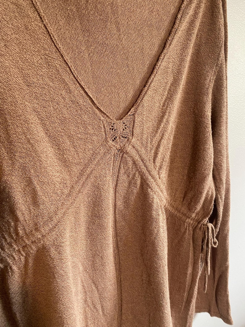 Puede incluir: Una blusa de punto marr&oacute;n con cuello en V y mangas largas. La blusa presenta un detalle calado decorativo en el escote y lazos en los laterales. La tela tiene un aspecto texturizado.