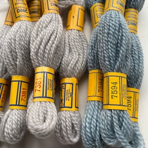 May include: Close-up of several skeins of DMC embroidery floss in shades of gray and blue. Each skein is labeled with a yellow and blue paper band, displaying the number and brand information. The image showcases the texture and color variations of the thread.